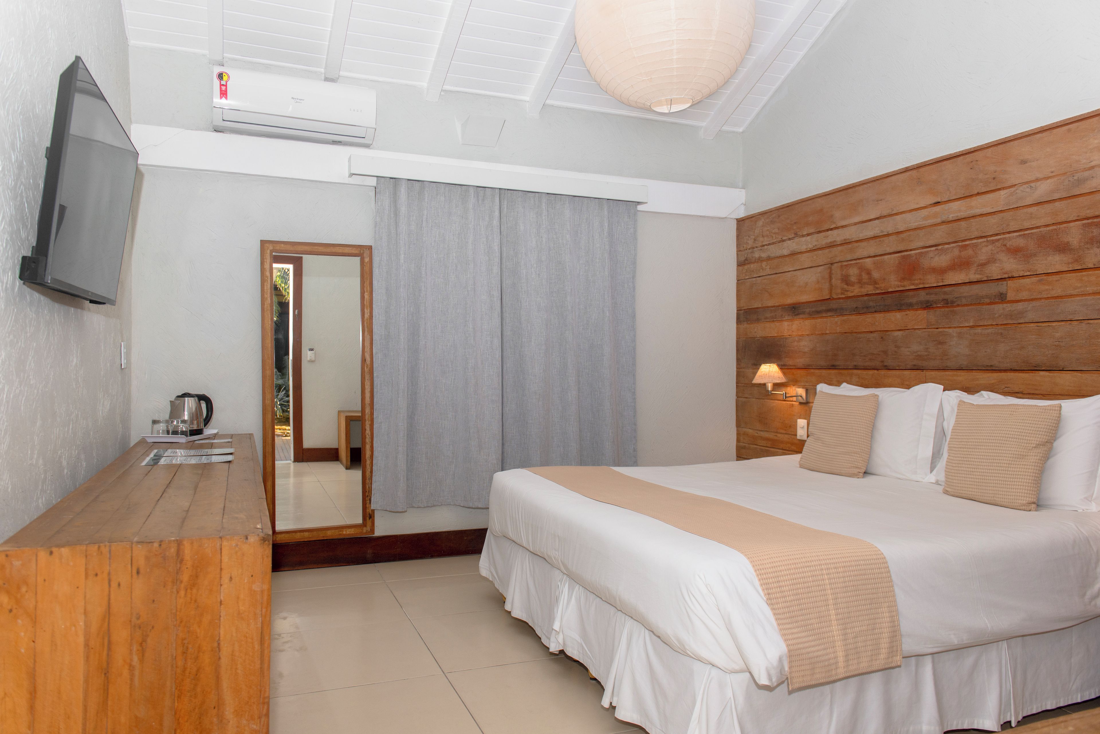 Ein Hotelzimmer mit einem hölzernen Kopfteil, einem Bett mit weißer Bettwäsche, einem Holztisch mit Wasserkocher, einem Flachbildfernseher und einem Deckenventilator.