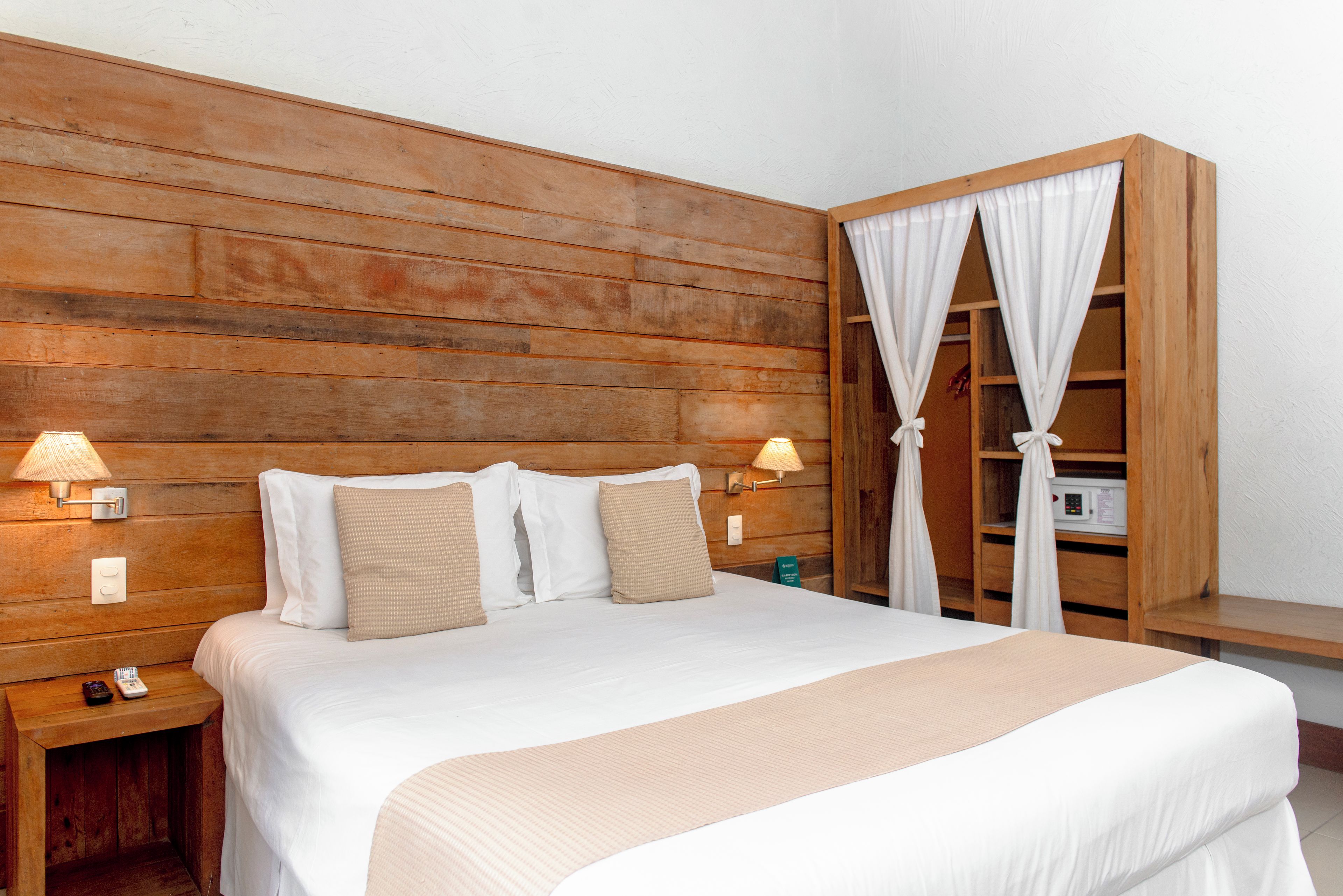 A hotel room with a wooden headboard, a bed with white linens, two bedside lamps, and a wooden nightstand with a remote control.