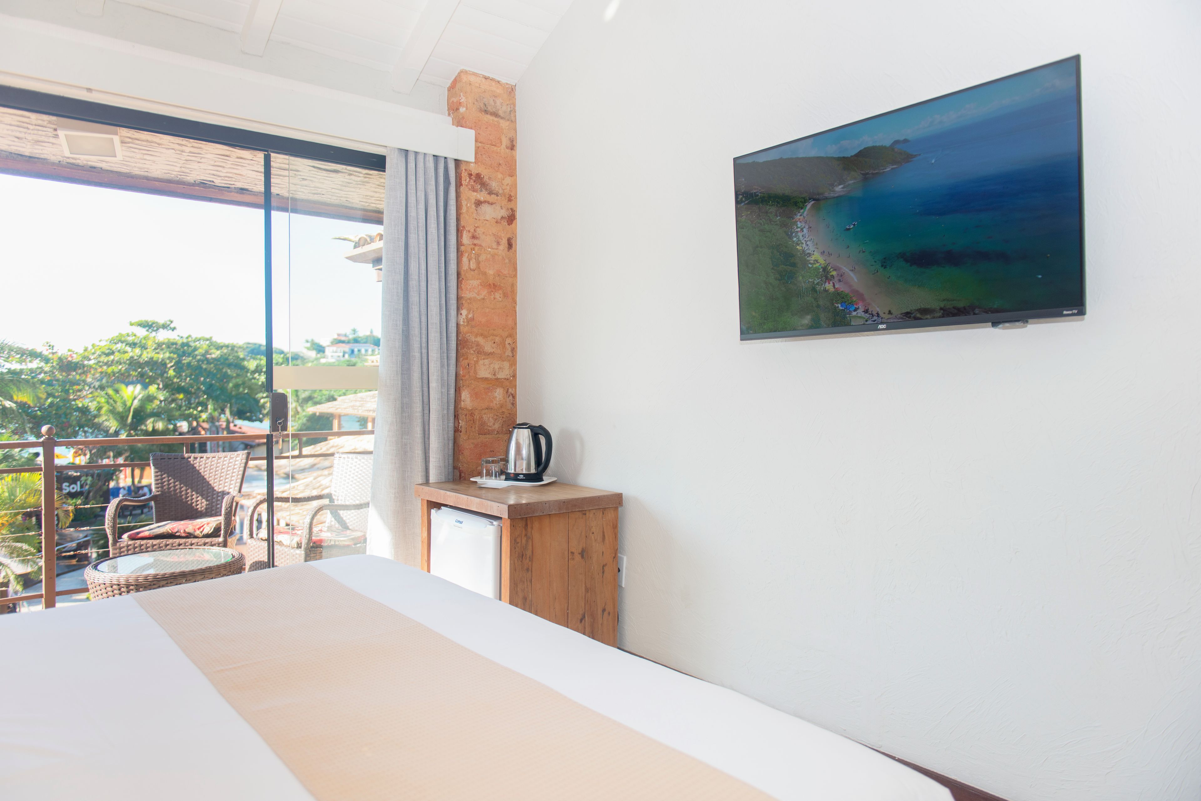 Une chambre d’hôtel avec un lit, une petite table en bois sur laquelle se trouve une bouilloire, un tableau représentant une plage et un balcon agrémenté de meubles en osier.