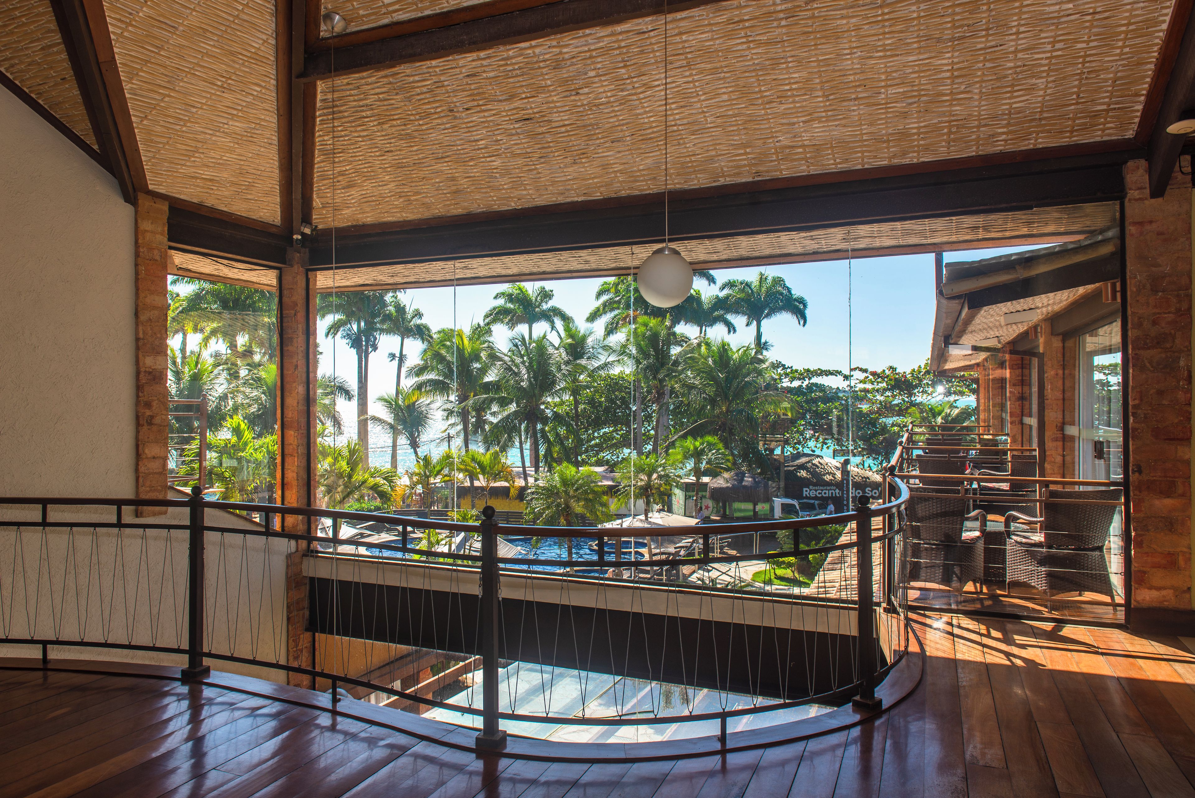 Un espacio interior amplio con suelo de madera, un balcón con vistas a palmeras y una piscina, y una lámpara colgante.