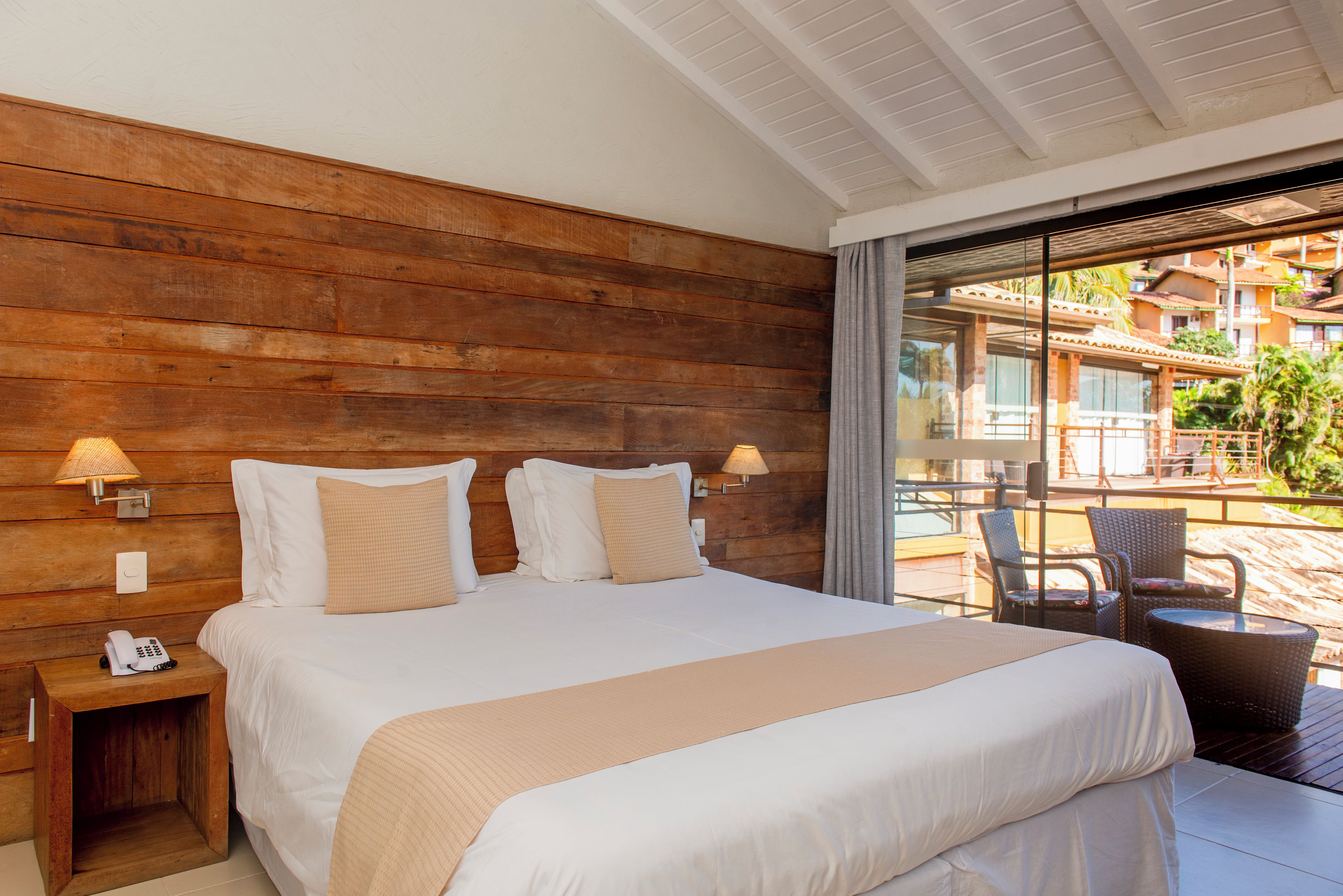 Een hotelkamer met een houten hoofdeinde, een groot bed met witte lakens, twee nachtlampjes en uitzicht op het buitengebied door een glazen deur.