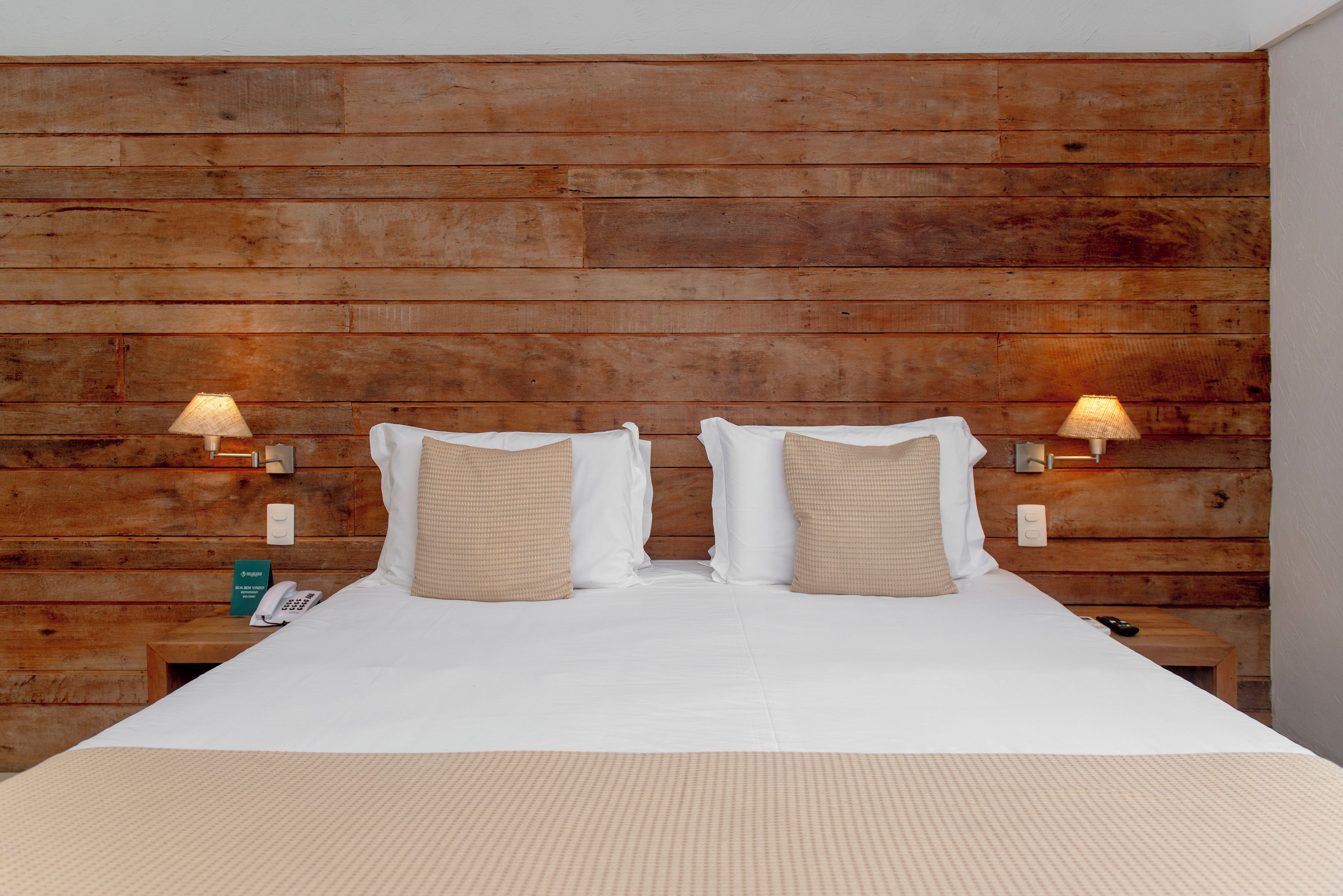 A hotel room with a wooden headboard, two bedside lamps, a bed with white linens, and a nightstand with a phone and a sign.