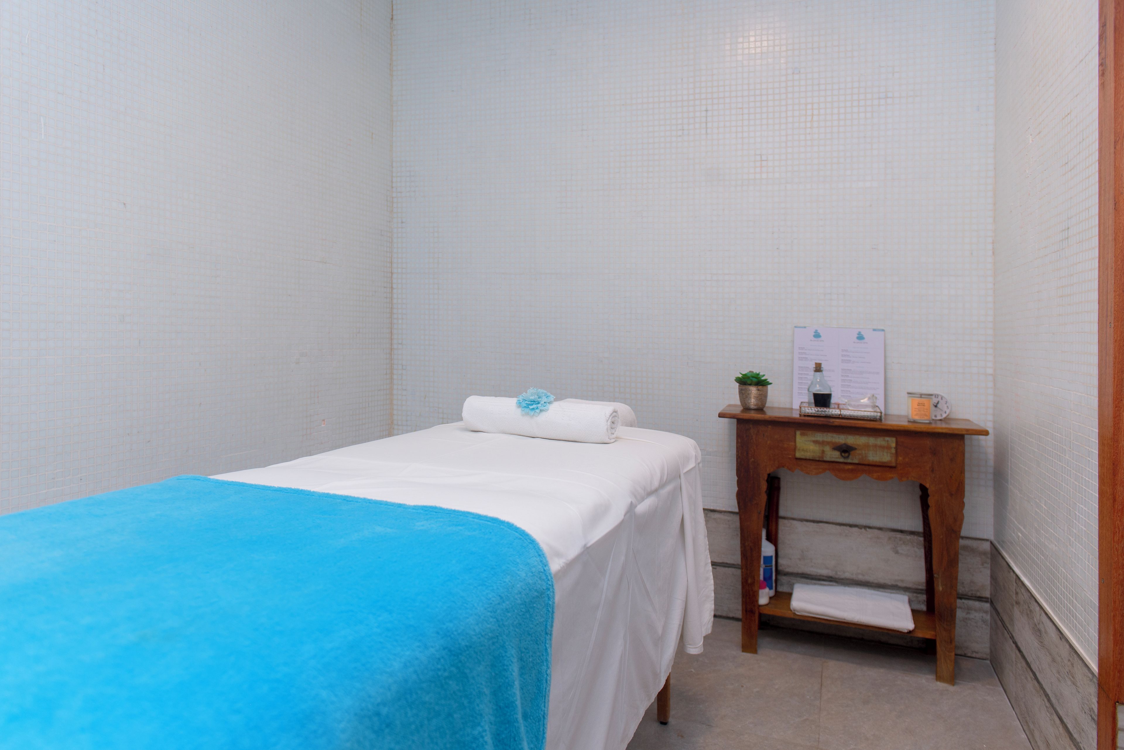 A small, clean room with a bed, a wooden side table, and a white tiled wall.