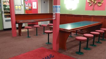 Uma sala de bilhar com mesa de sinuca verde, banquetas vermelhas e tapete vermelho com os dizeres "Bienvenue" e "K. PALACE".