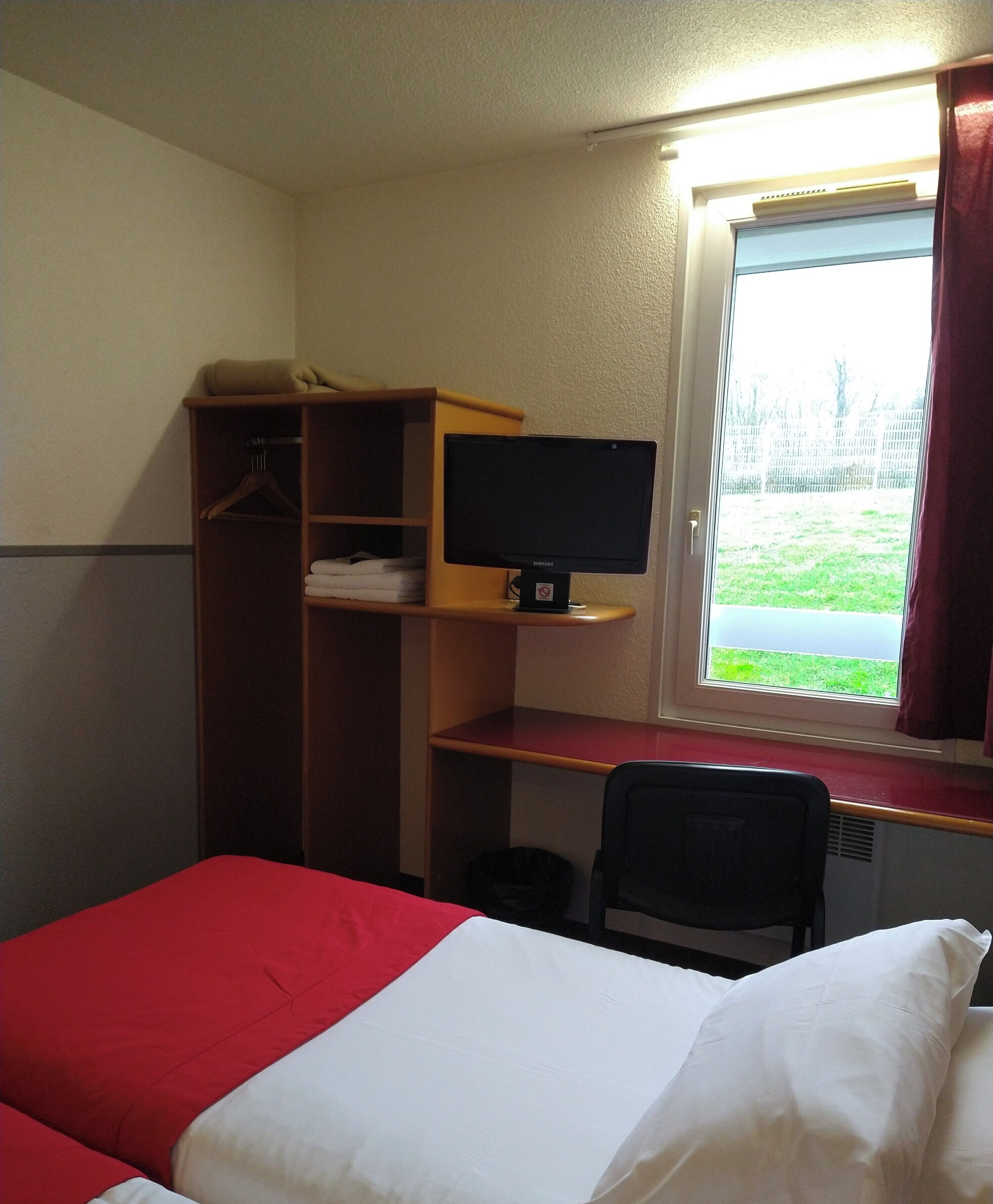 A hotel room with a bed, a desk with a computer, a chair, a wardrobe, and a window with a view of the outdoors.