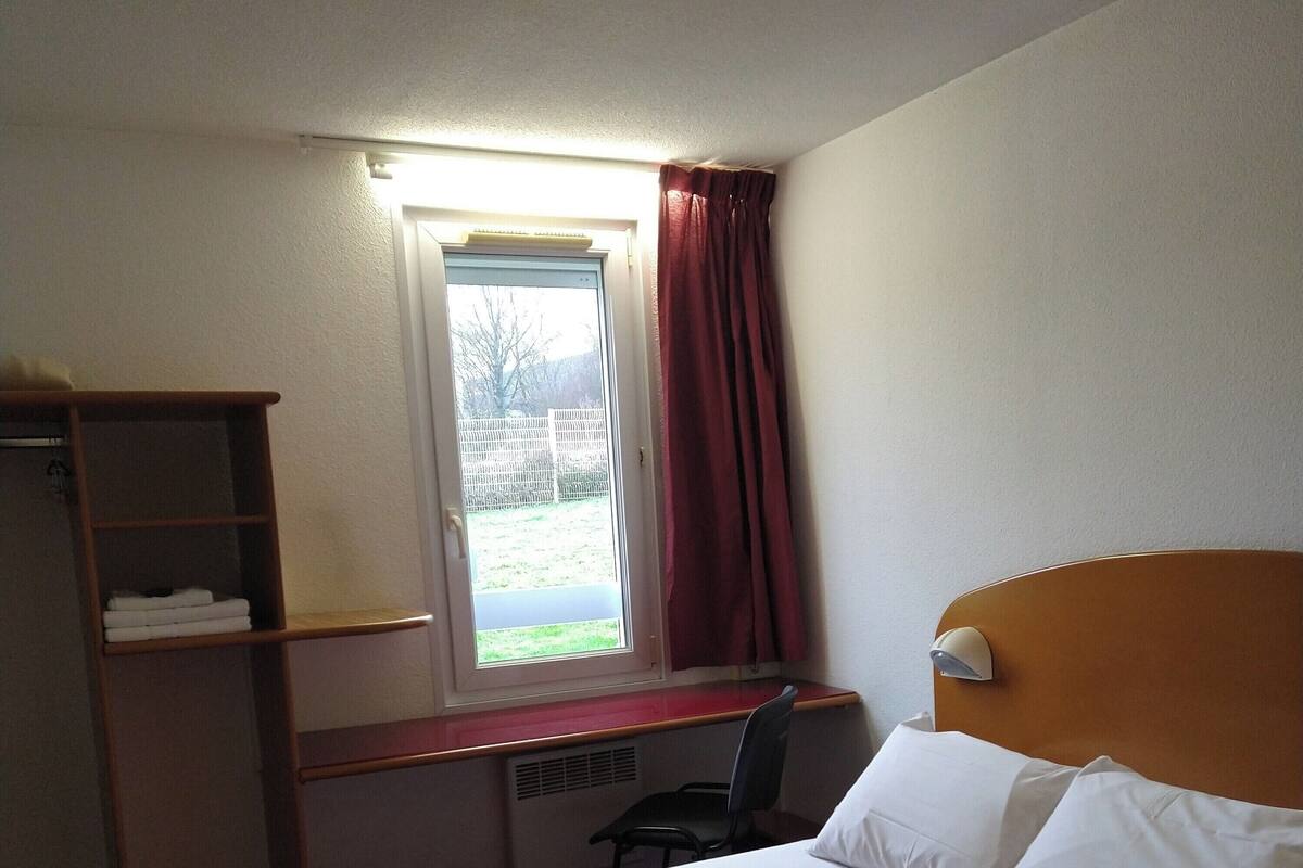 Une chambre d’hôtel comprenant un lit, un bureau avec une chaise, une fenêtre avec des rideaux et une étagère avec des serviettes pliées.
