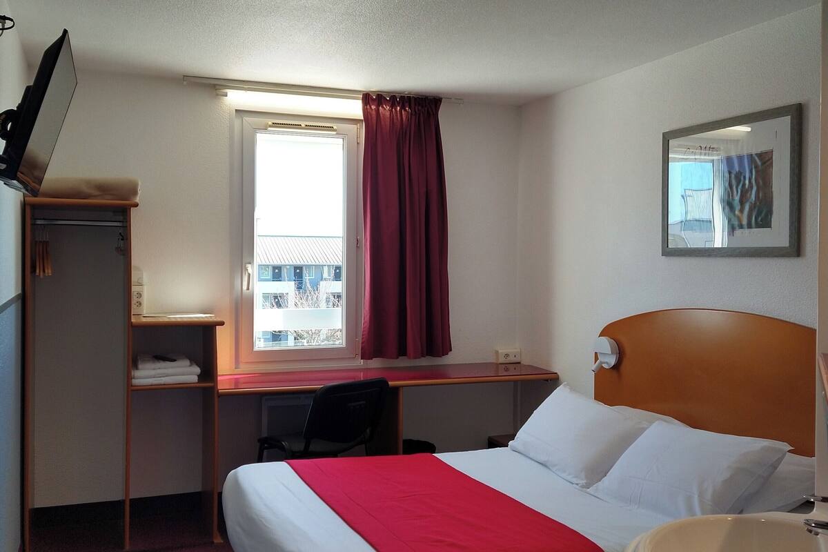 Une chambre d’hôtel comprenant un lit, un bureau, une chaise, une télévision, une fenêtre avec des rideaux, une salle de bain avec un lavabo et un tableau encadré au mur.