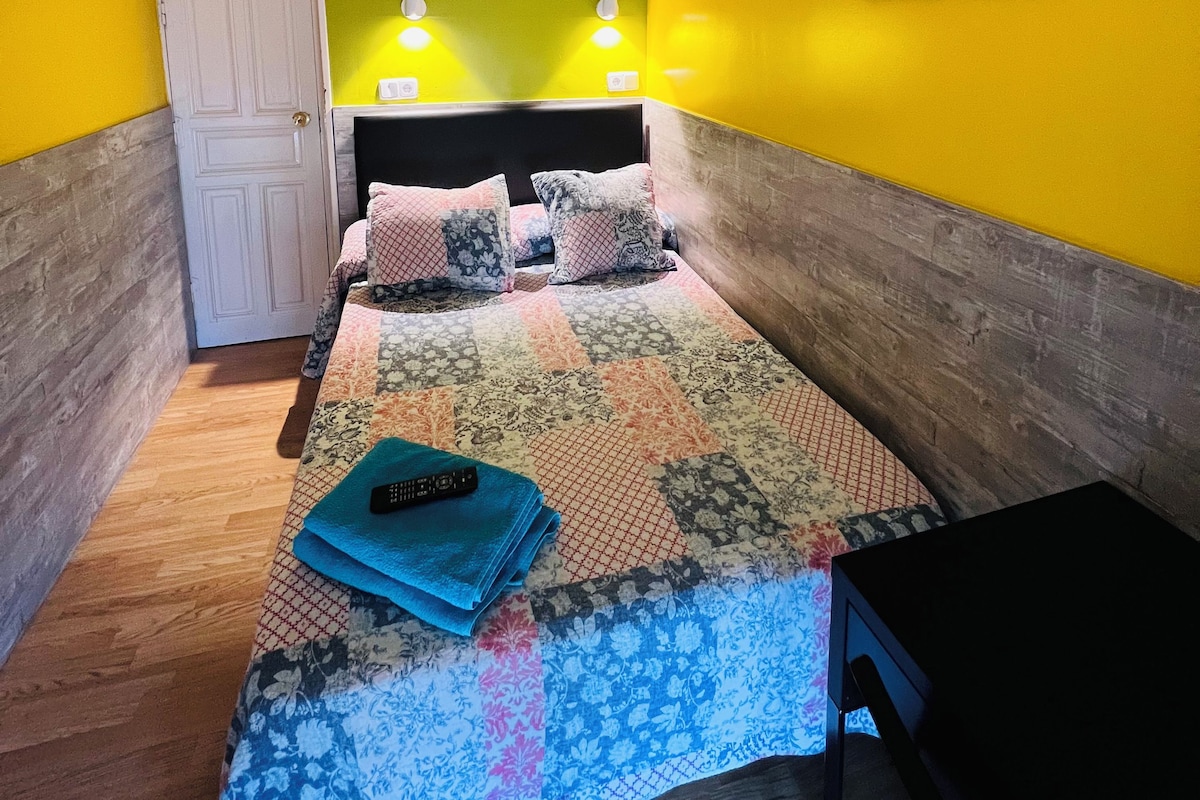 Um quarto pequeno e colorido com uma cama, duas luminárias de cabeceira, uma porta e um quadro de um veado na parede.