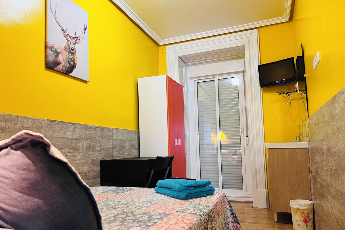Quarto com cama, escrivaninha, cadeira, televisão e porta que dá para uma varanda.