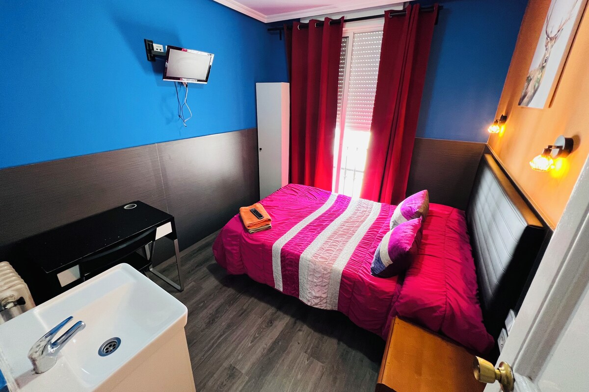 Une chambre d’hôtel de taille réduite, comprenant un lit, un bureau, une chaise, une petite table, un lavabo et un téléviseur fixé au mur.