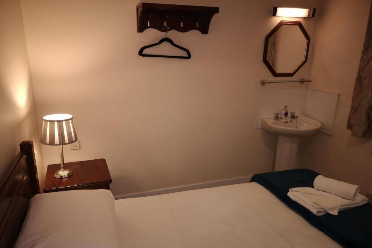 A hotel room with a bed, a bedside table with a lamp, a hanging coat rack, a sink, and a mirror.