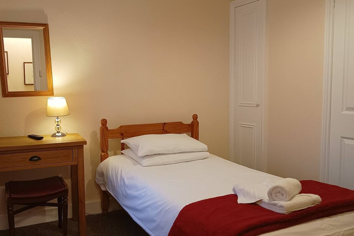 A neatly made bed with white linens and a red blanket, a wooden nightstand with a lamp, a small wooden chair, and a mirror on the wall.