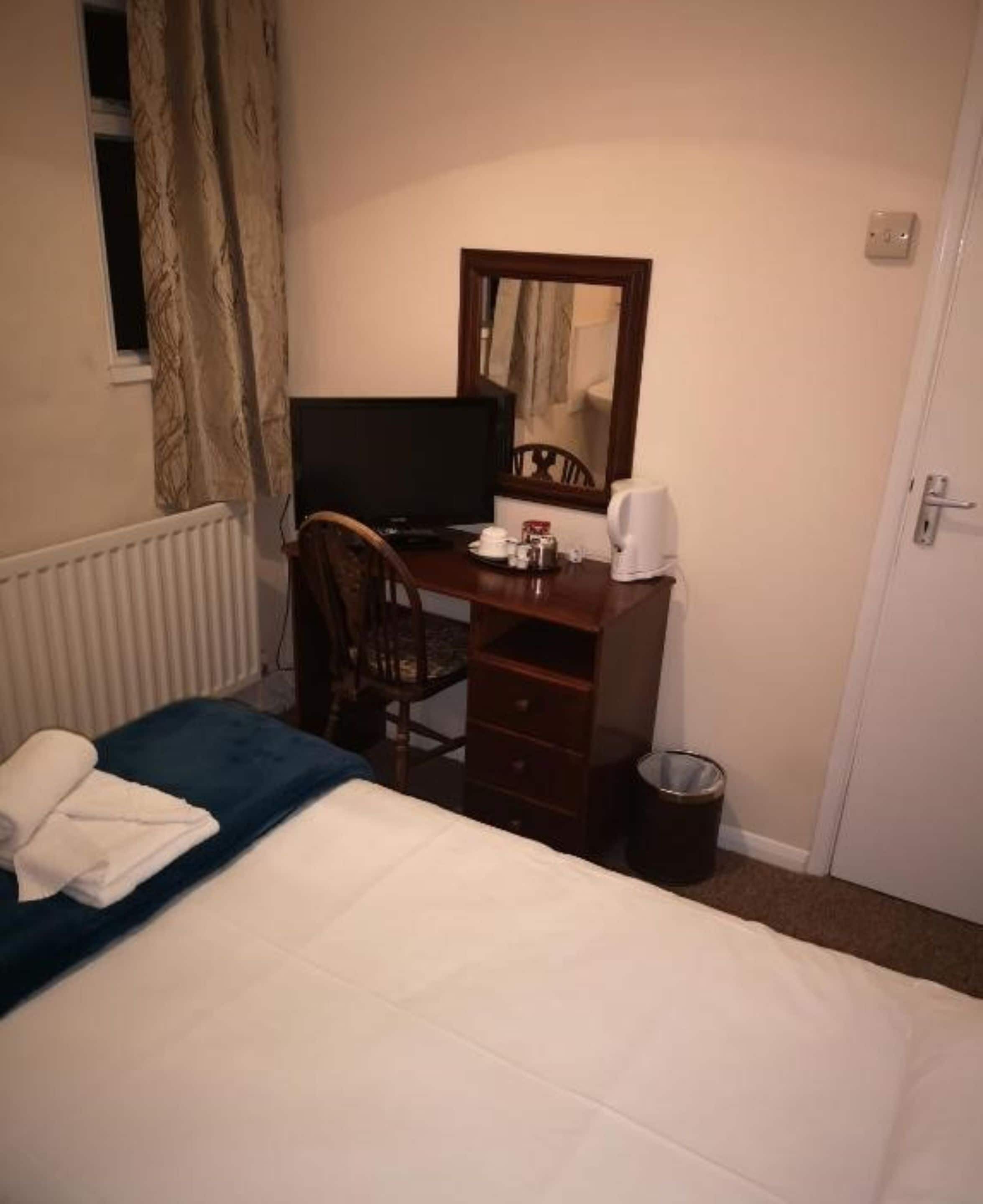 A hotel room with a bed, a desk with a computer, a chair, a small trash bin, and a window with curtains.