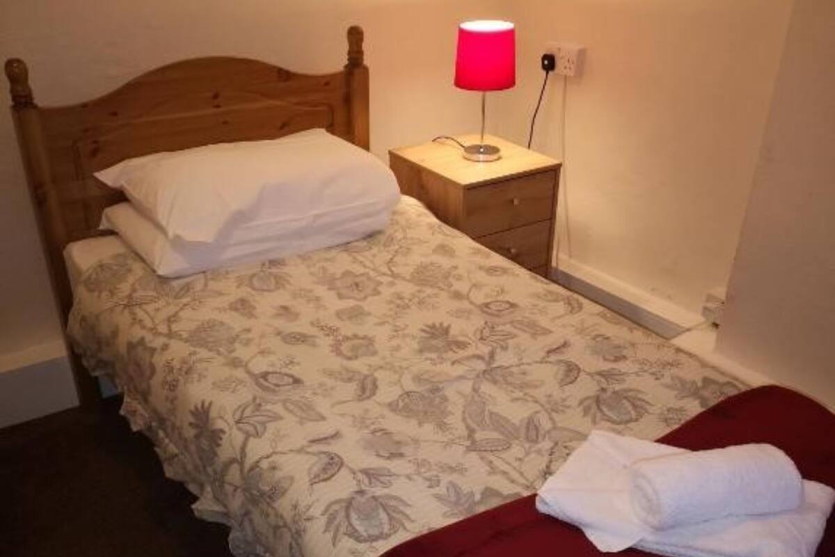 A single bed with a floral bedspread, a wooden headboard, a nightstand with a red lamp, and a red throw blanket.
