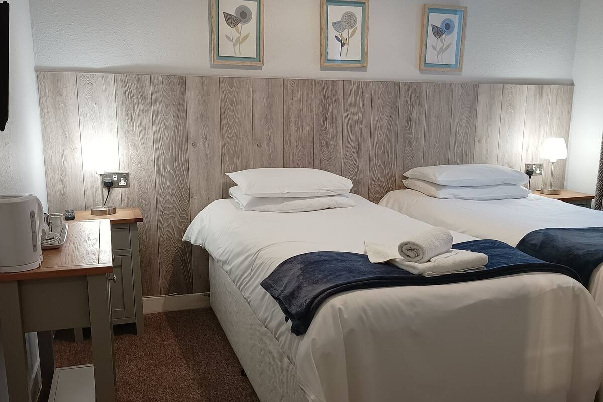 A hotel room with two beds, a wooden headboard, a desk with a lamp, and artwork on the wall.