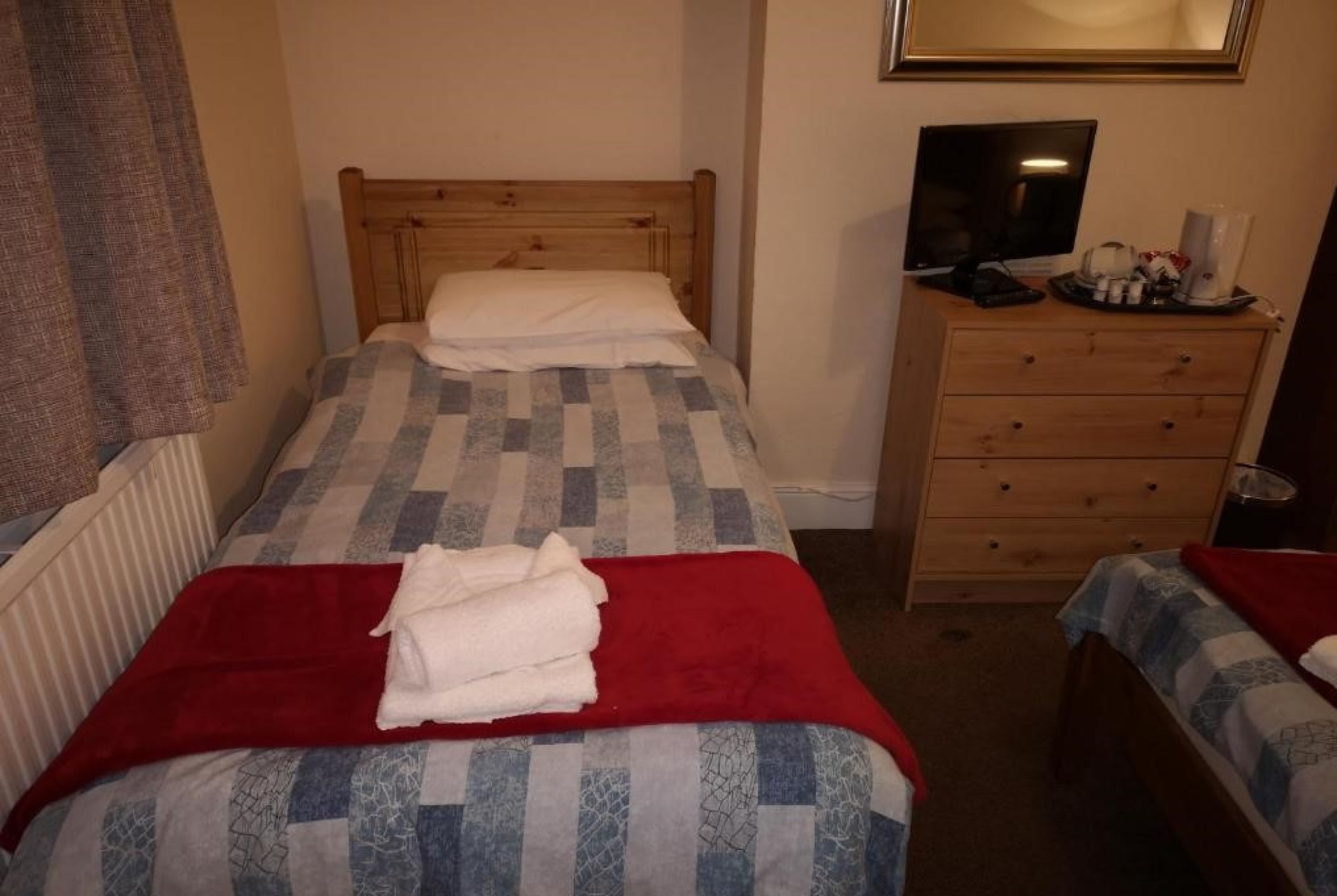 A small, neatly arranged bedroom with two single beds, a wooden dresser, a television, and a mirror.