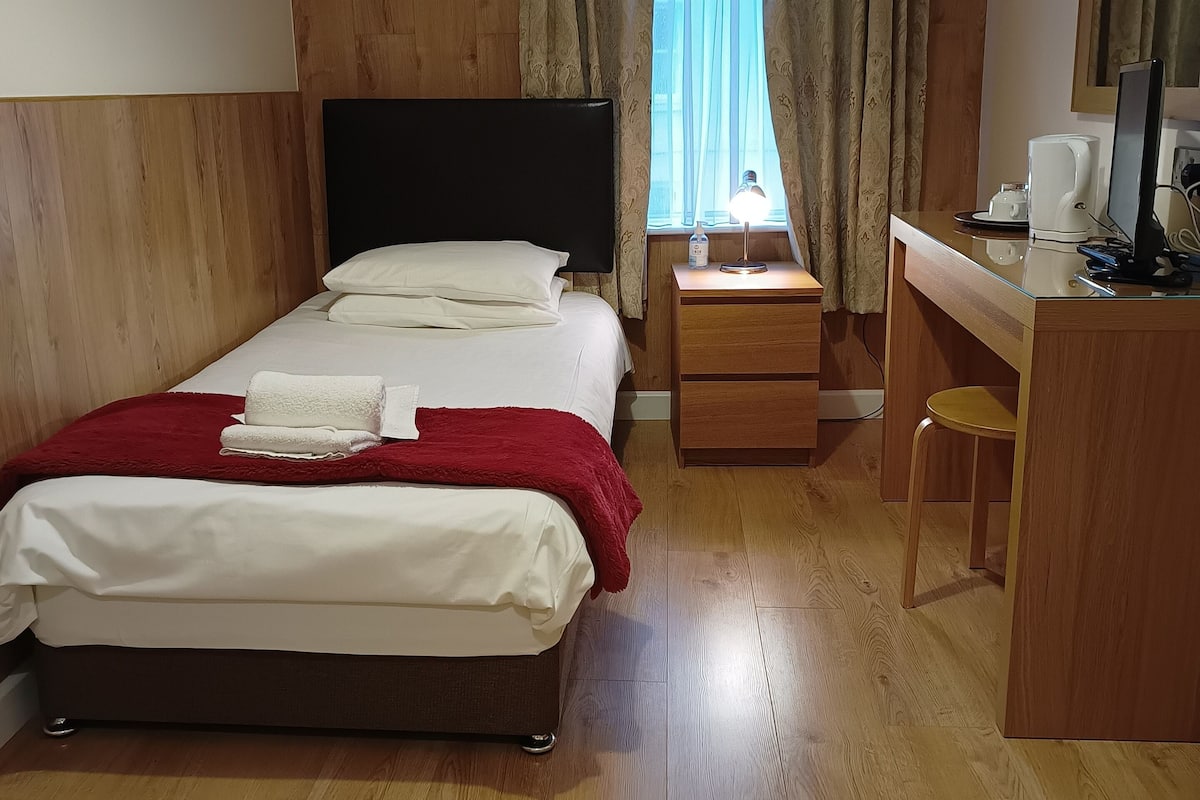 A hotel room with a bed, a desk, a chair, a window with curtains, and a bedside lamp.