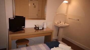 A small, compact hotel room with a single bed, a desk with a television, a bedside table, a mirror, and a sink.