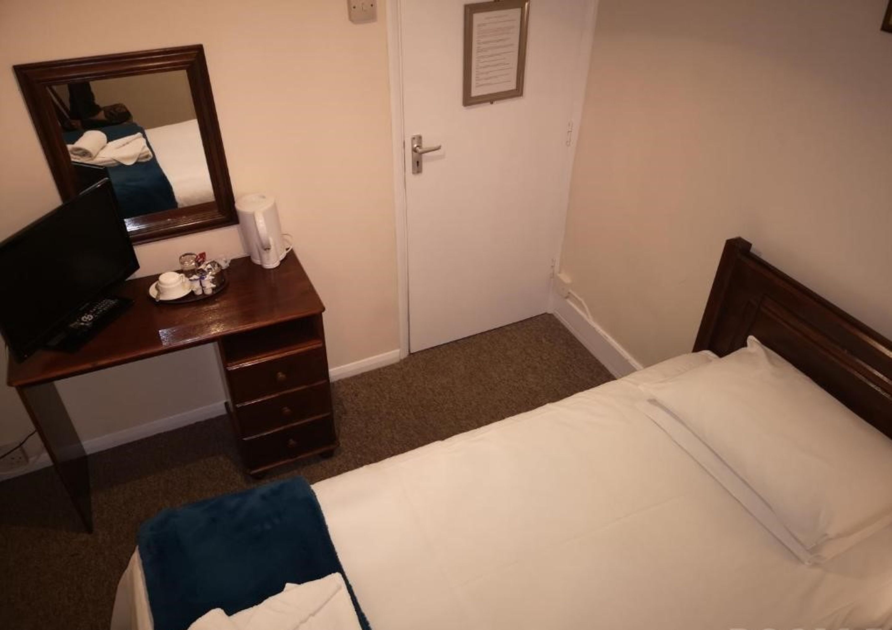 Quarto de hotel com uma cama de solteiro, uma cômoda de madeira, um espelho e uma porta com uma placa.