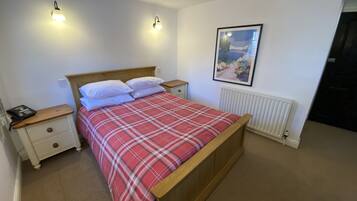 Quarto com cama de madeira, mesinhas de cabeceira brancas, colcha xadrez vermelha, duas luminárias na parede, radiador branco e um quadro emoldurado.