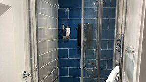 A glass-enclosed shower with a blue-tiled wall, a handheld showerhead, and a towel rack with towels. - The White Hart (Newport Pagnell)