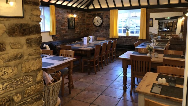 A rustic pub interior with exposed brick walls, wooden tables, and a sloped ceiling with exposed beams. - The White Hart (Newport Pagnell)