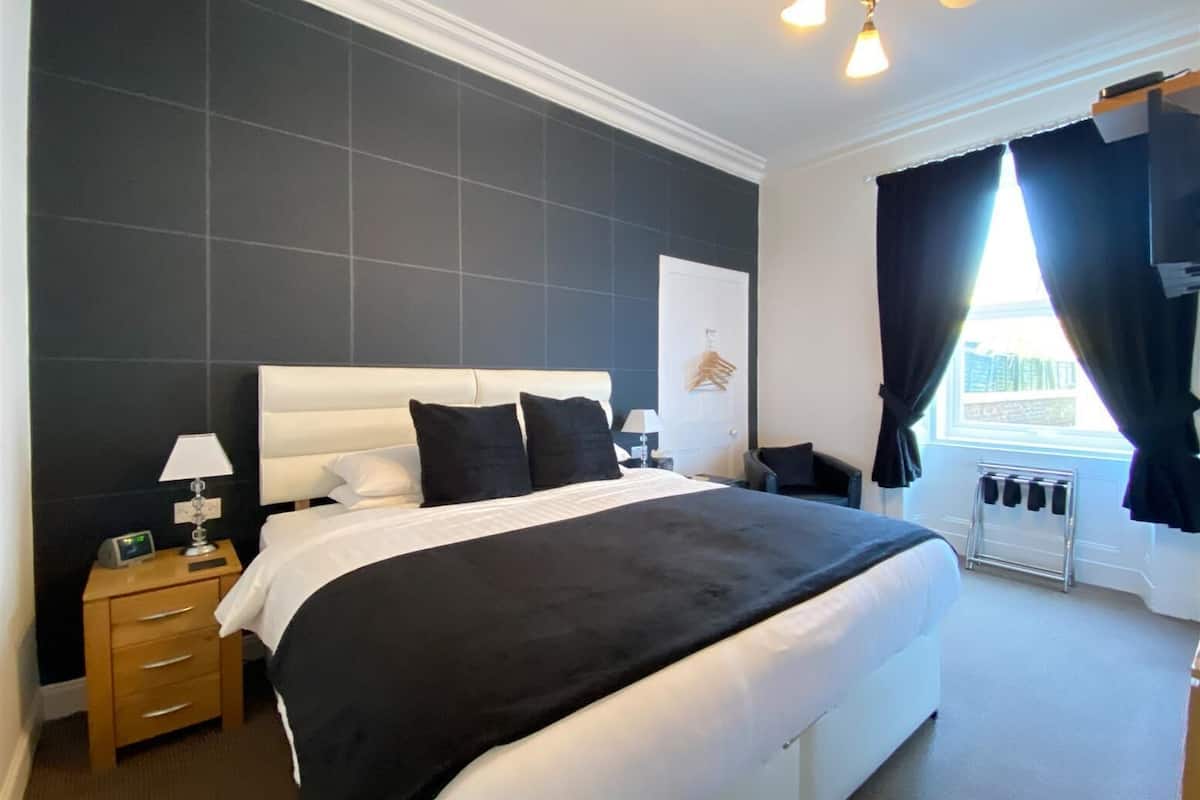 A modern hotel room with a large bed, bedside tables, a nightstand, a lamp, a radiator, and a window with curtains.