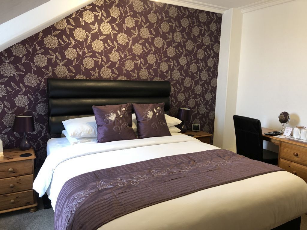 A bedroom with a large bed, a dark headboard, and a floral-patterned wallpaper. There are two bedside tables with lamps and a desk with a chair.