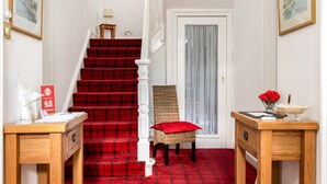 A staircase with red carpet leading to a door with a white curtain. There are two wooden side tables with a vase of red flowers and a bowl of lemons. - Rosebank Guest House (Perth)