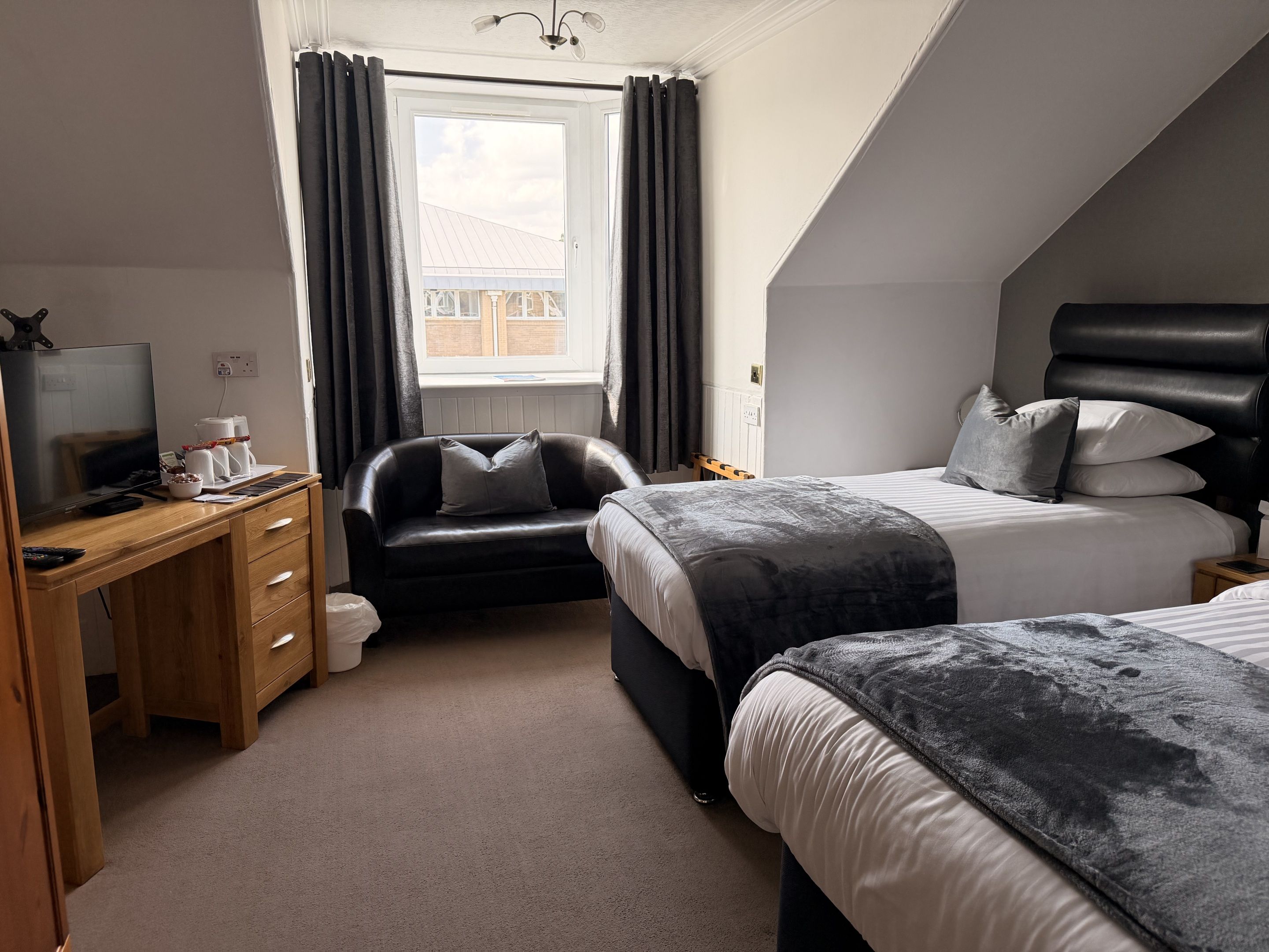 A bedroom with a desk, two beds, a window with curtains, and a view of the outside.