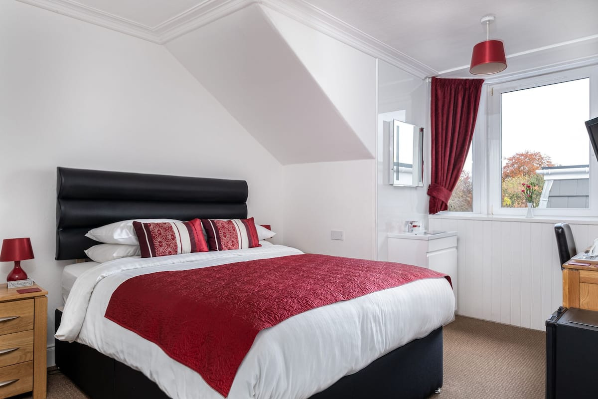 A bedroom with a black headboard, white bedding, a red accent throw, a wooden nightstand with a red lamp, a desk with a computer, and a window with a red curtain.