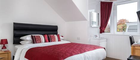 Un dormitorio con cabecera negra, ropa de cama blanca, un plaid rojo decorativo, una mesita de noche de madera con una lámpara roja, un escritorio con una computadora y una ventana con cortina roja.