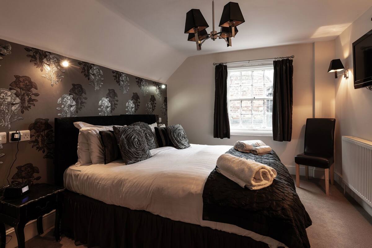 A bedroom with a large bed, a headboard, a window with curtains, a wall-mounted TV, and a radiator.