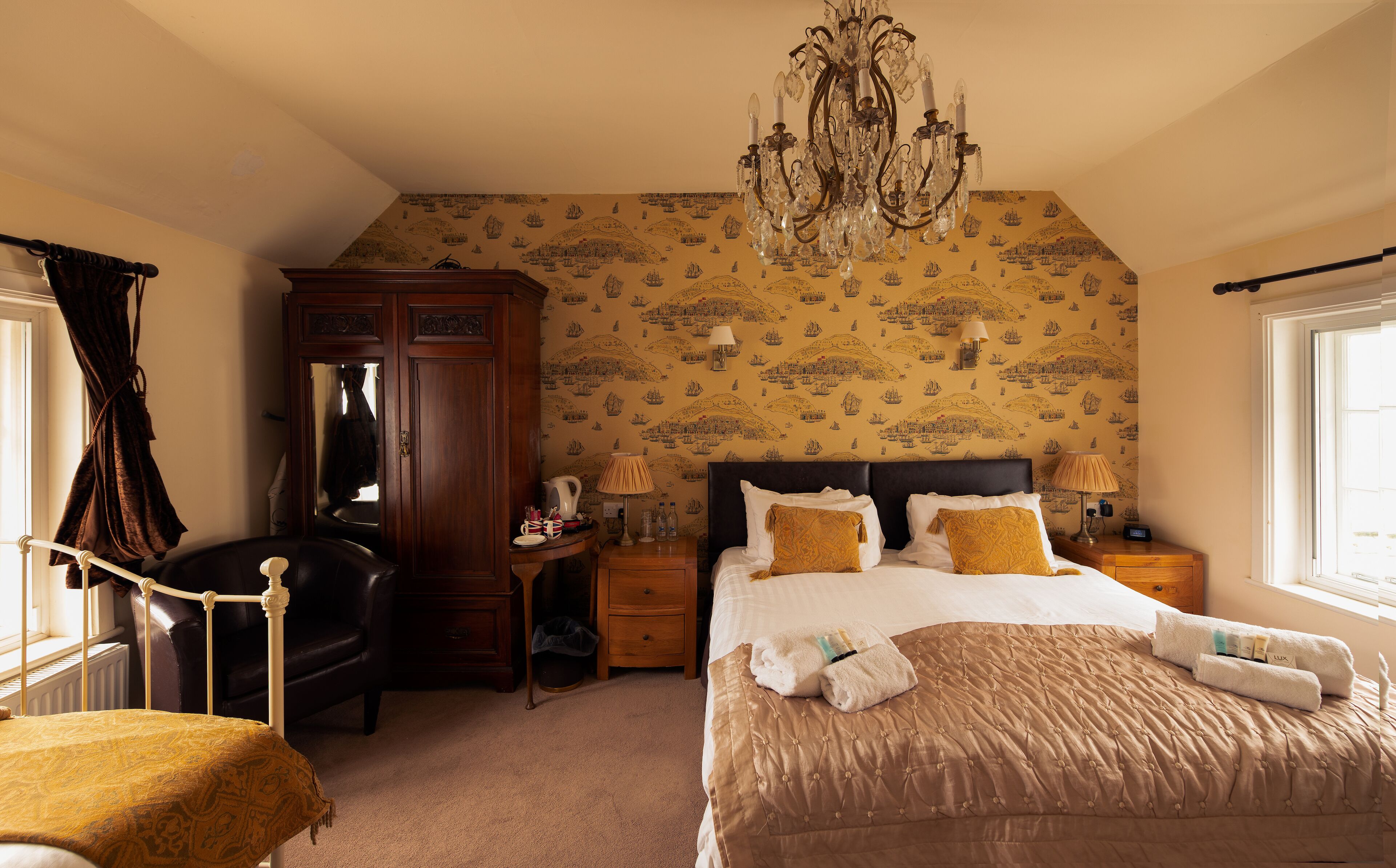 A bedroom with a large bed, a wooden wardrobe, a desk with a lamp, and a chandelier.