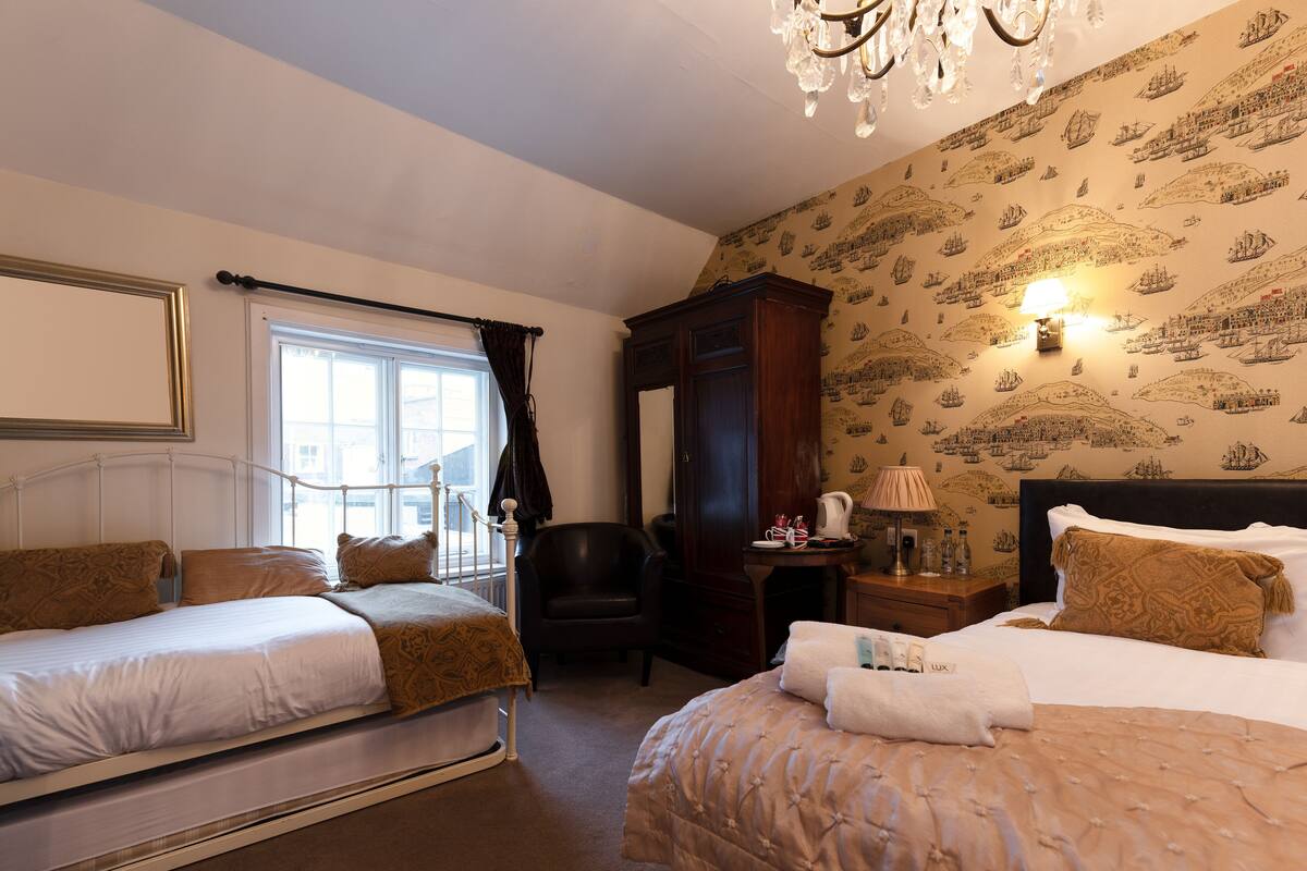 A hotel room with two beds, a sitting area, a chandelier, and a wall with maritime-themed wallpaper.