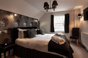 A bedroom with a large bed, a nightstand, a chair, a window with curtains, a wall-mounted TV, and a ceiling light fixture.