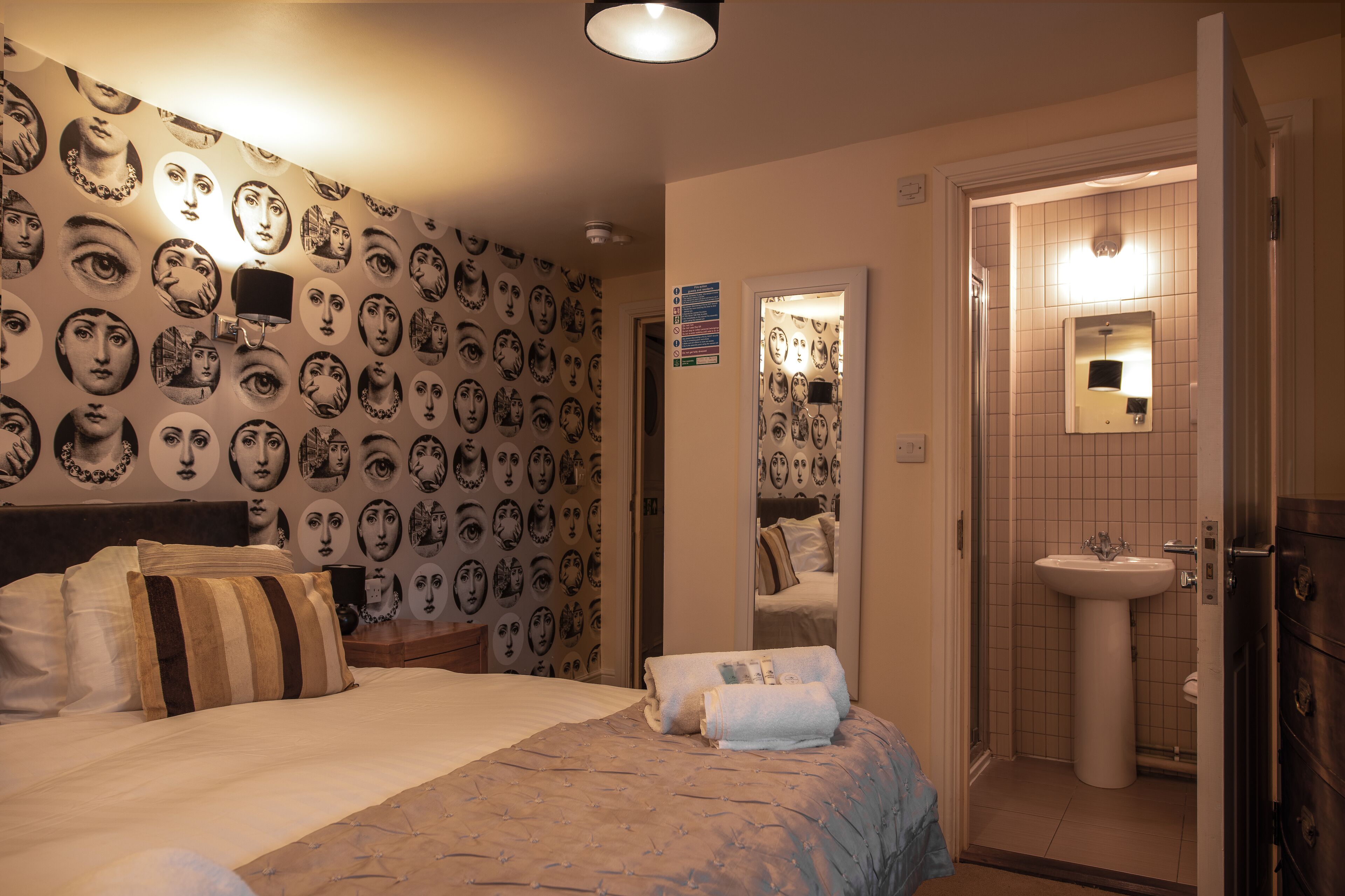 A hotel room with a patterned wall, a bed with striped pillows, a bedside table, and a bathroom with a sink and mirror.