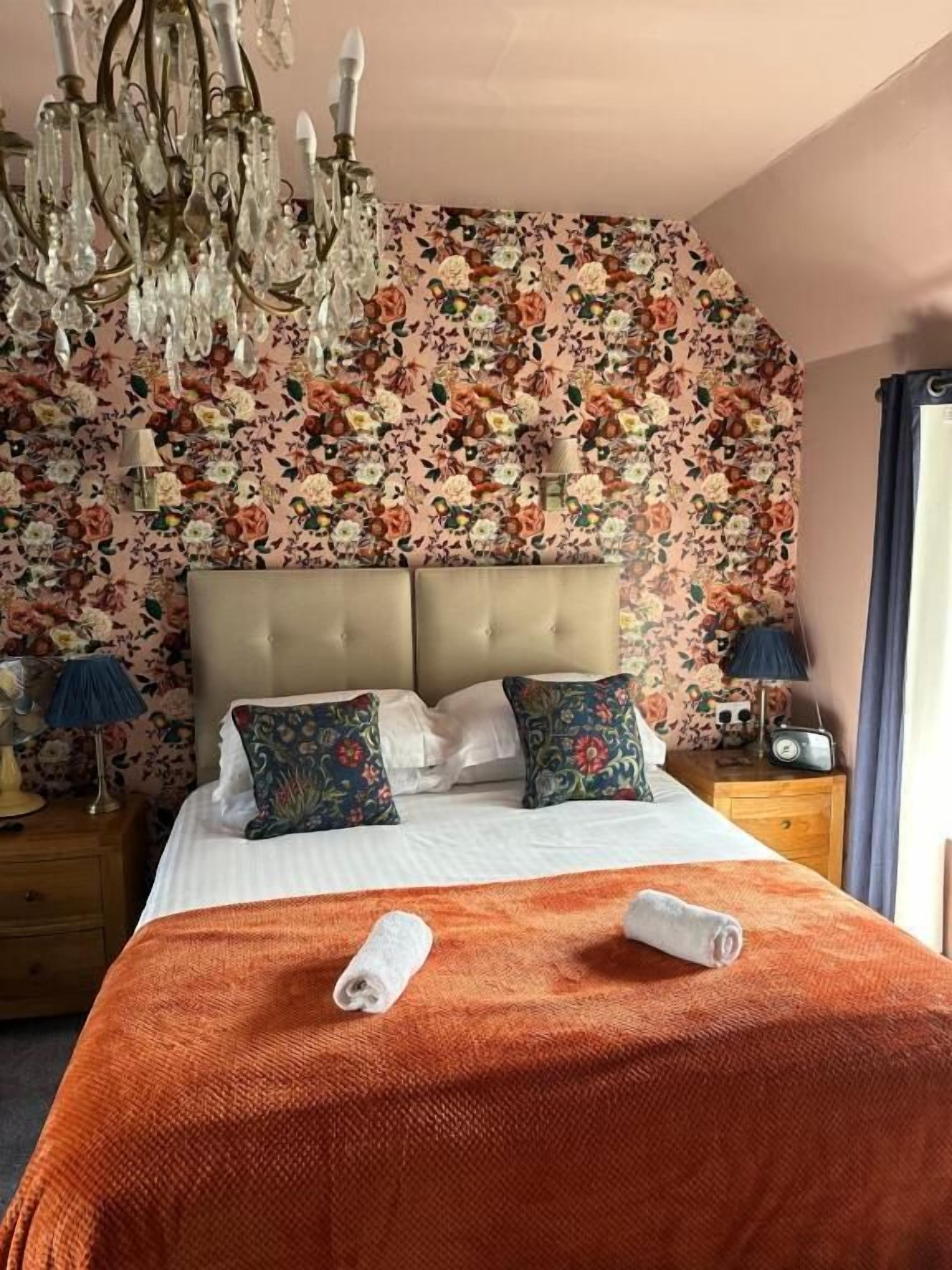 A hotel room with a floral wallpaper, a tufted headboard, a bed with an orange blanket, two rolled towels, a chandelier, bedside lamps, and a radio.