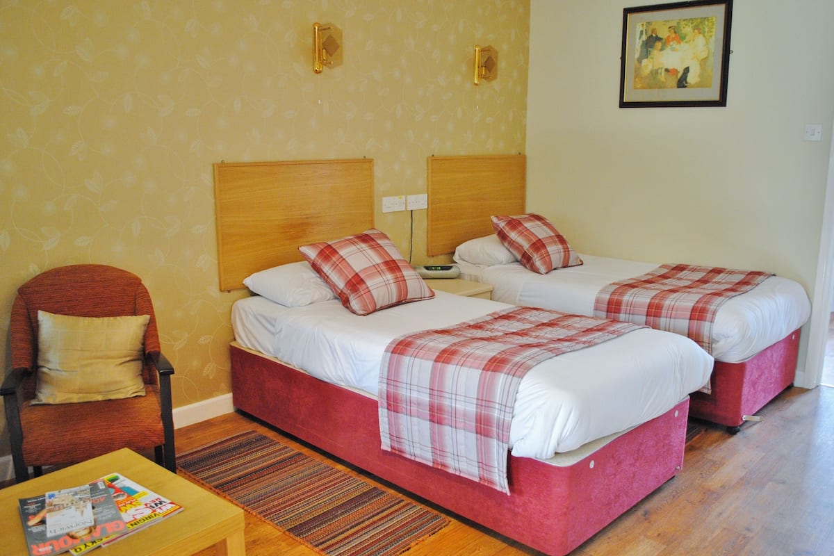 Habitación de hotel con dos camas, una silla, una mesita con revistas y un cuadro en la pared.