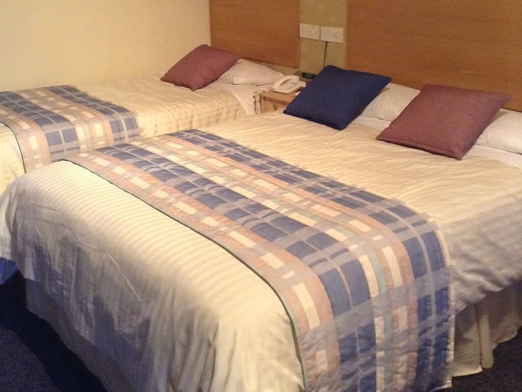 A hotel room with two beds, each with a patterned bedspread and pillows. There is a bedside table with a telephone and a lamp.