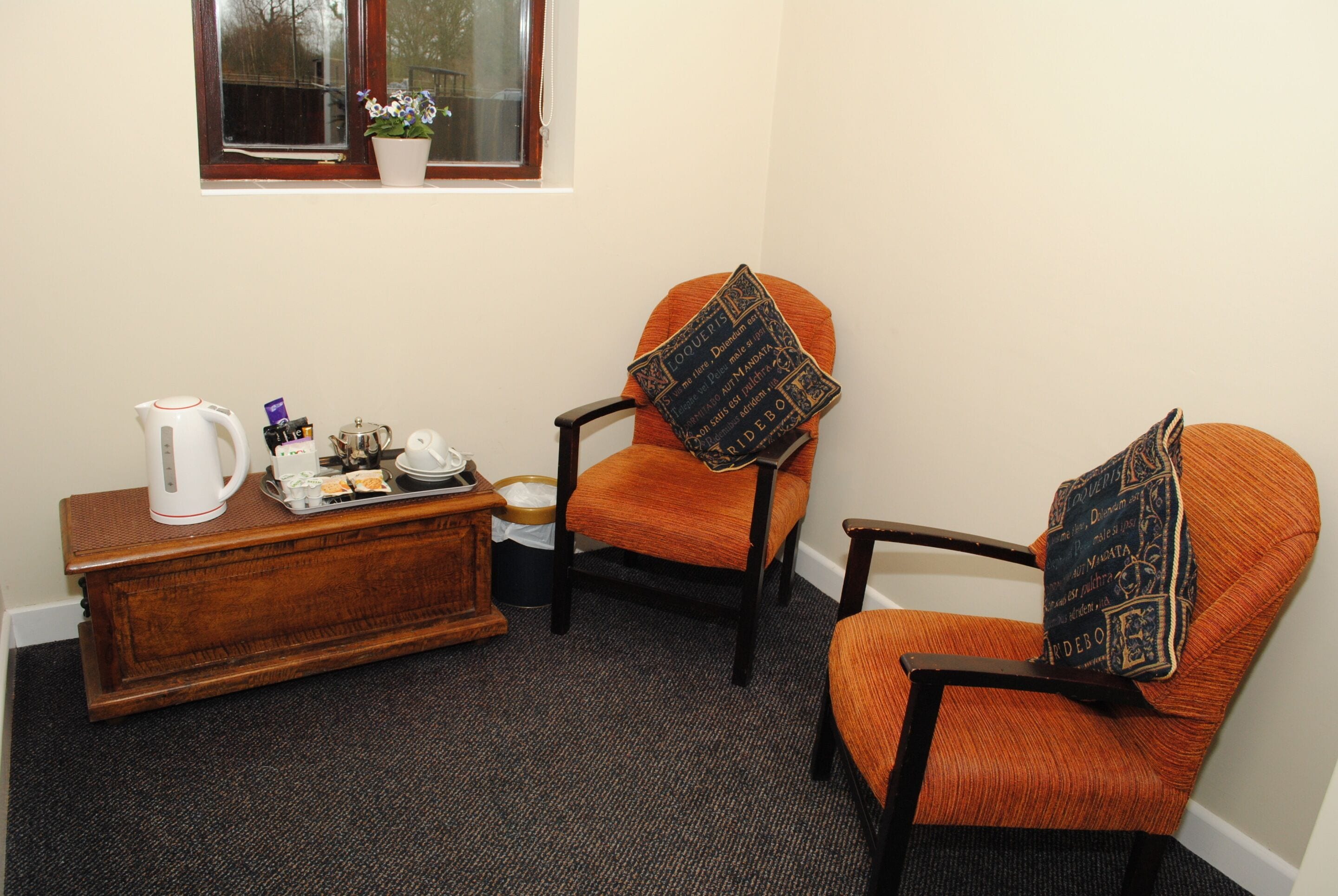 Una habitación con un aparador de madera, dos sillones tapizados en color naranja y cojines estampados, una ventana con planta en maceta y suelo alfombrado.