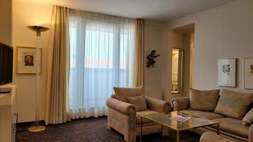 Una sala de estar con sofá, sillón y una mesa de centro de vidrio. Cuenta con un ventanal con cortinas traslúcidas, una lámpara de pie y cuadros enmarcados en las paredes.