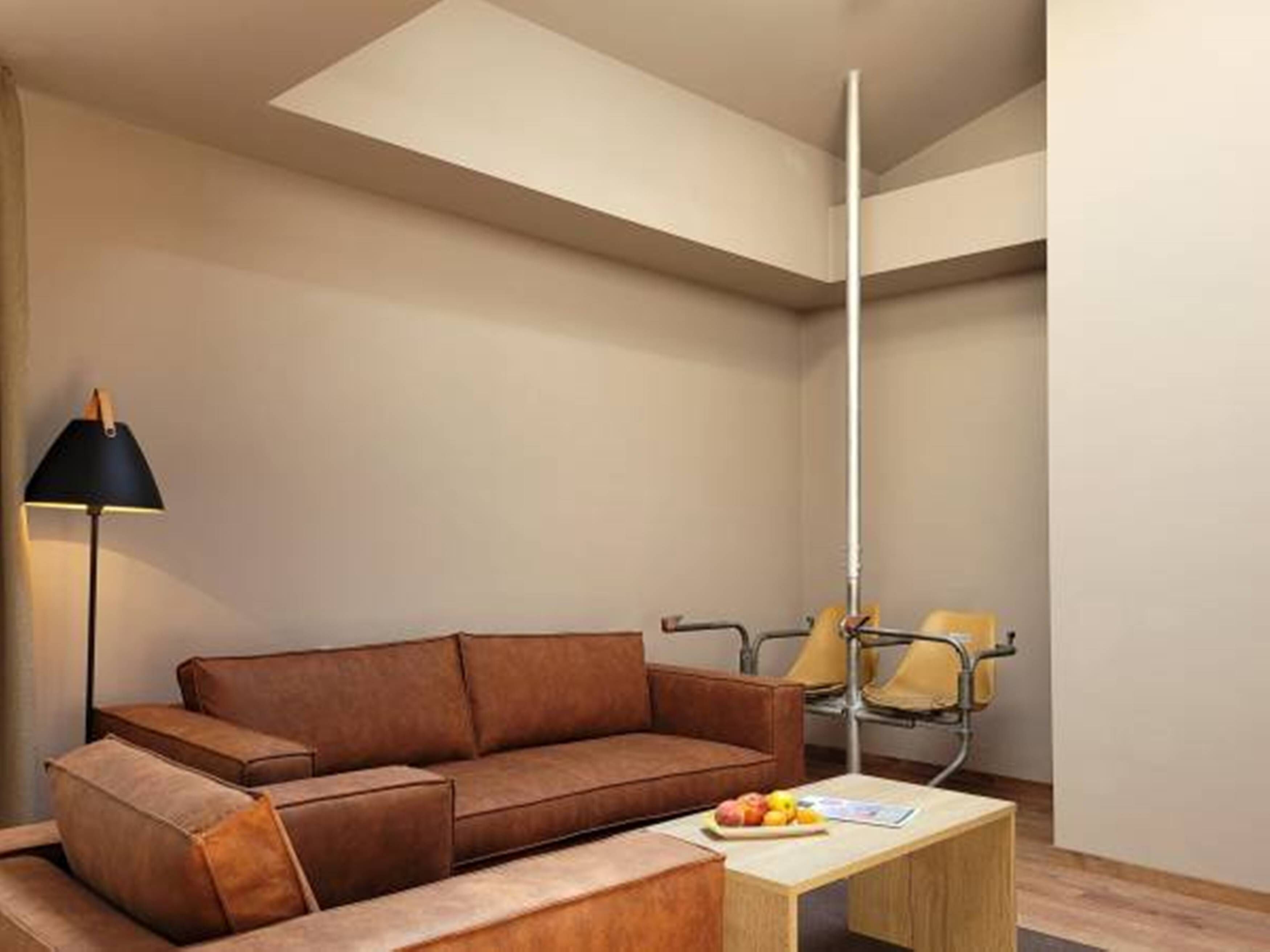 Ein modernes Wohnzimmer mit einem braunen Ledersofa, einem kleinen Holztisch mit Früchten, einer schwarzen Stehlampe und einem Sessel mit gelbem Kissen.