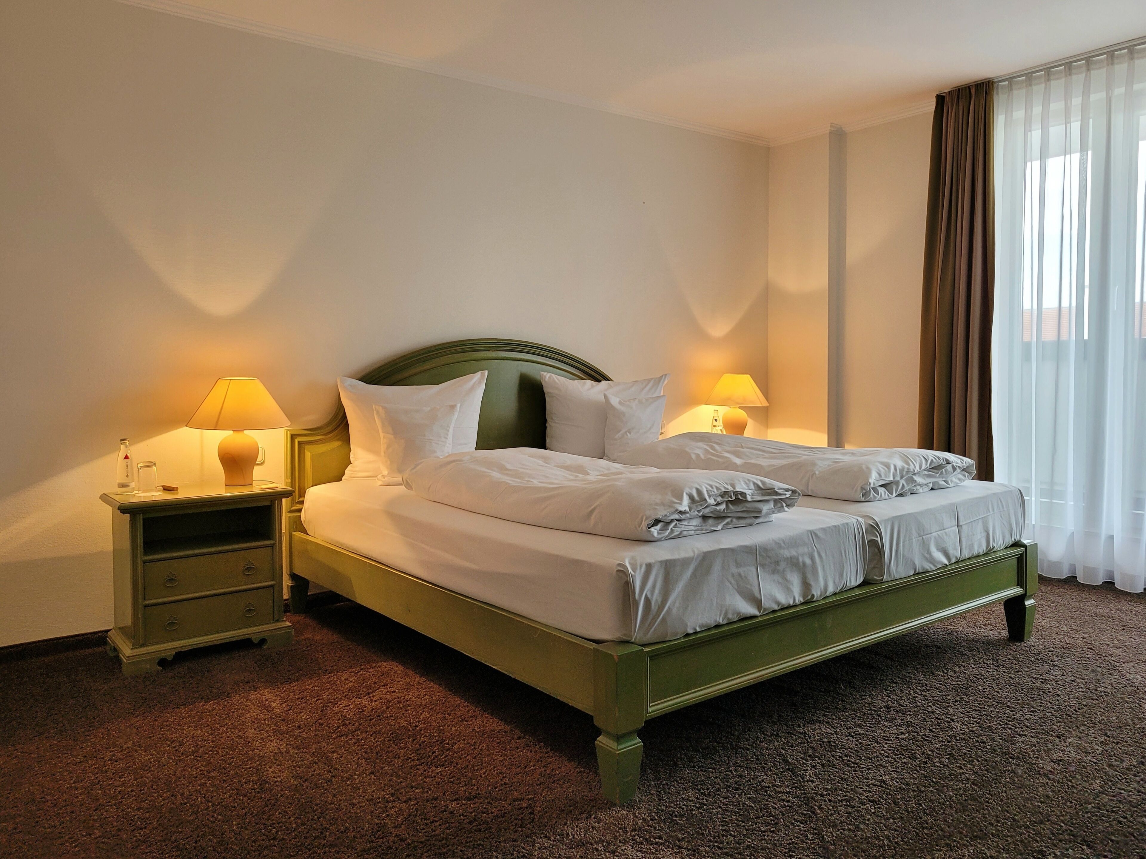 Una cama bien hecha con sábanas y almohadas blancas, dos mesitas de noche con lámparas y una ventana con cortinas translúcidas.