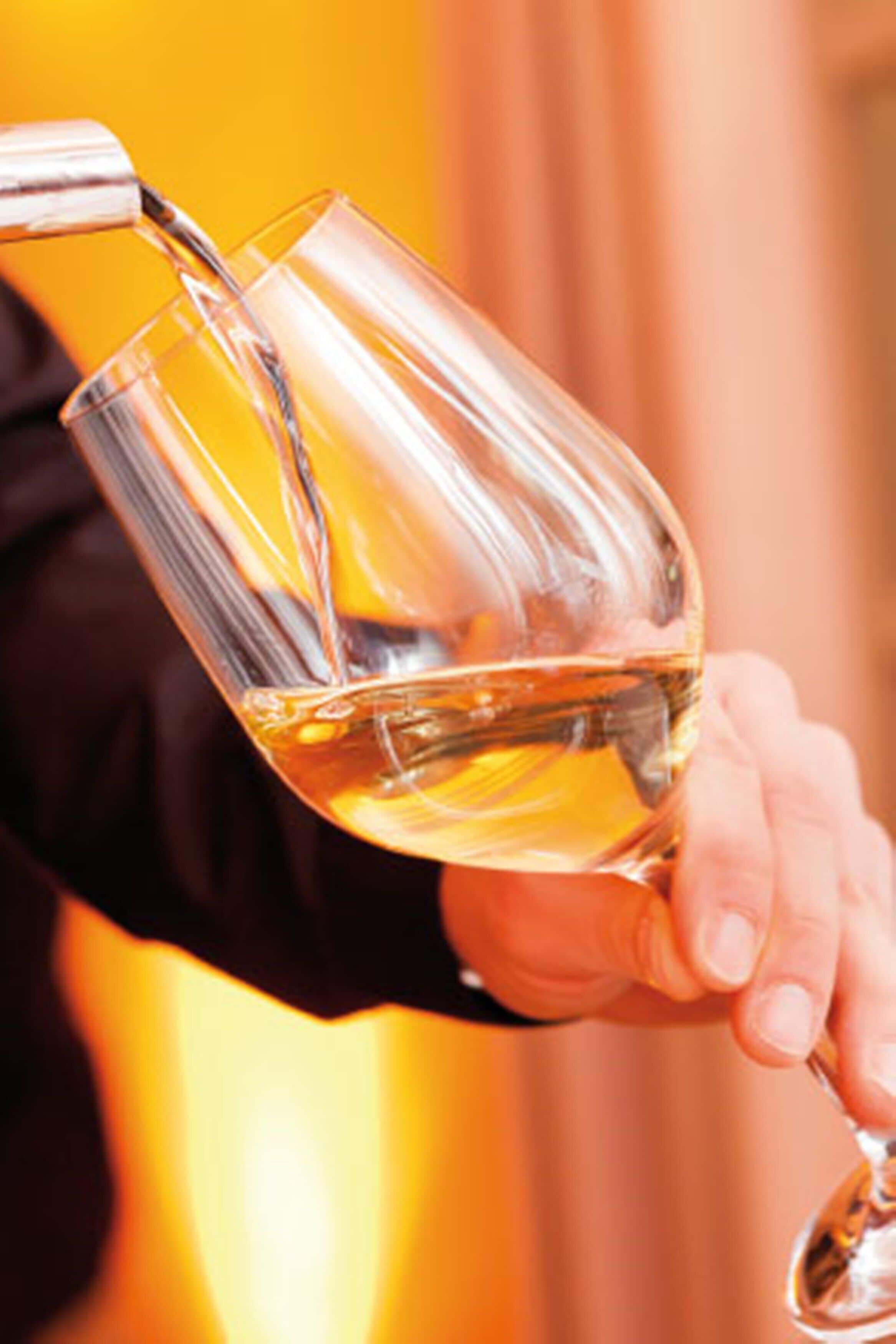 A hand holding a wine glass with a golden liquid being poured into it.