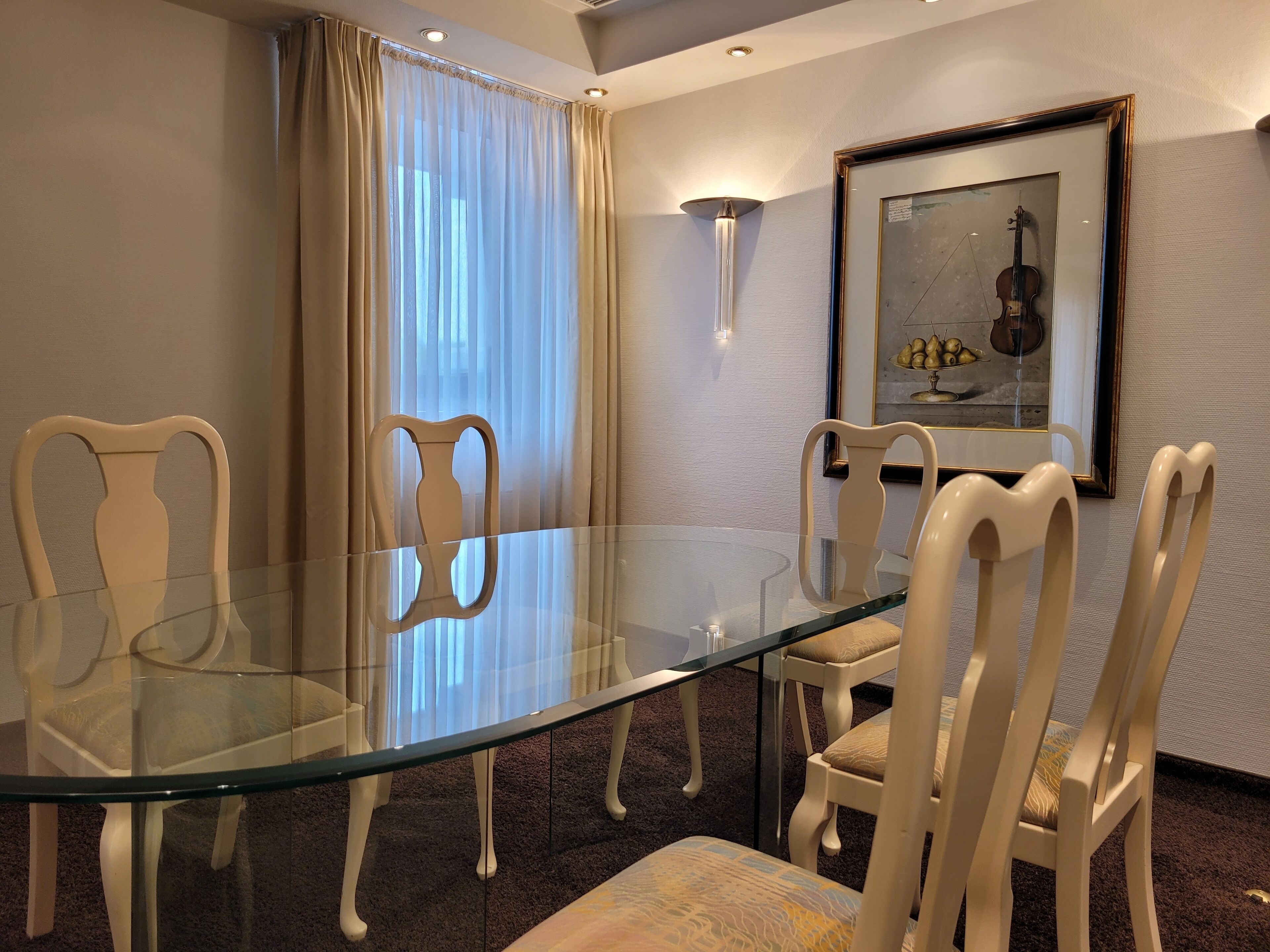 A conference room with a glass table and white chairs, a framed picture of a violin on the wall, and a window with sheer curtains.