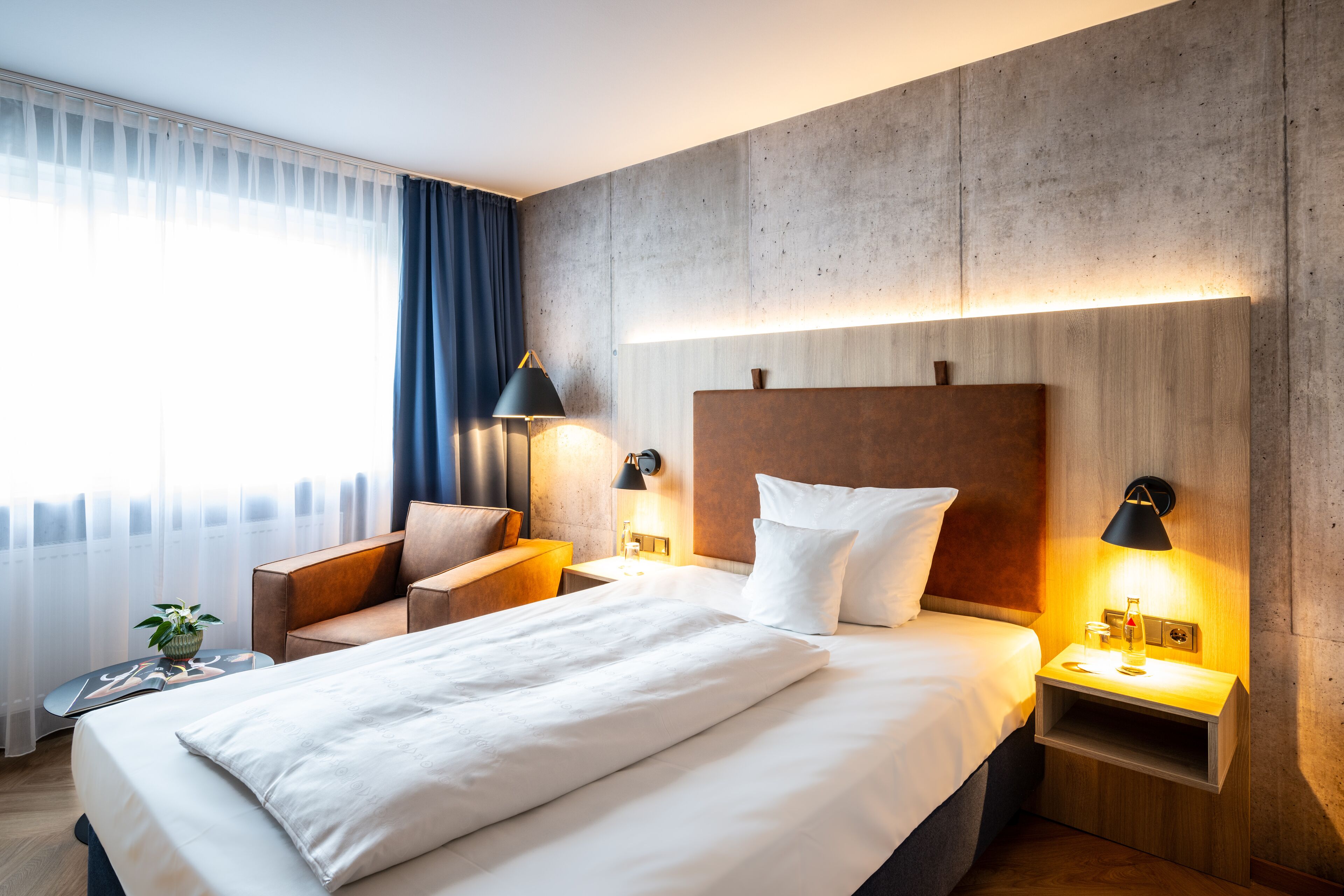 Una habitación de hotel moderna con una cama grande, un cabecero de madera, una mesita de noche con lámpara, una zona de estar con una silla y una pequeña mesa, y un ventanal con cortinas translúcidas.