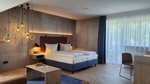 Una habitación de hotel moderna con una cama grande, un escritorio y una silla. Hay una mesita de centro y una mesita pequeña con una lámpara. La habitación tiene un ventanal con cortinas.