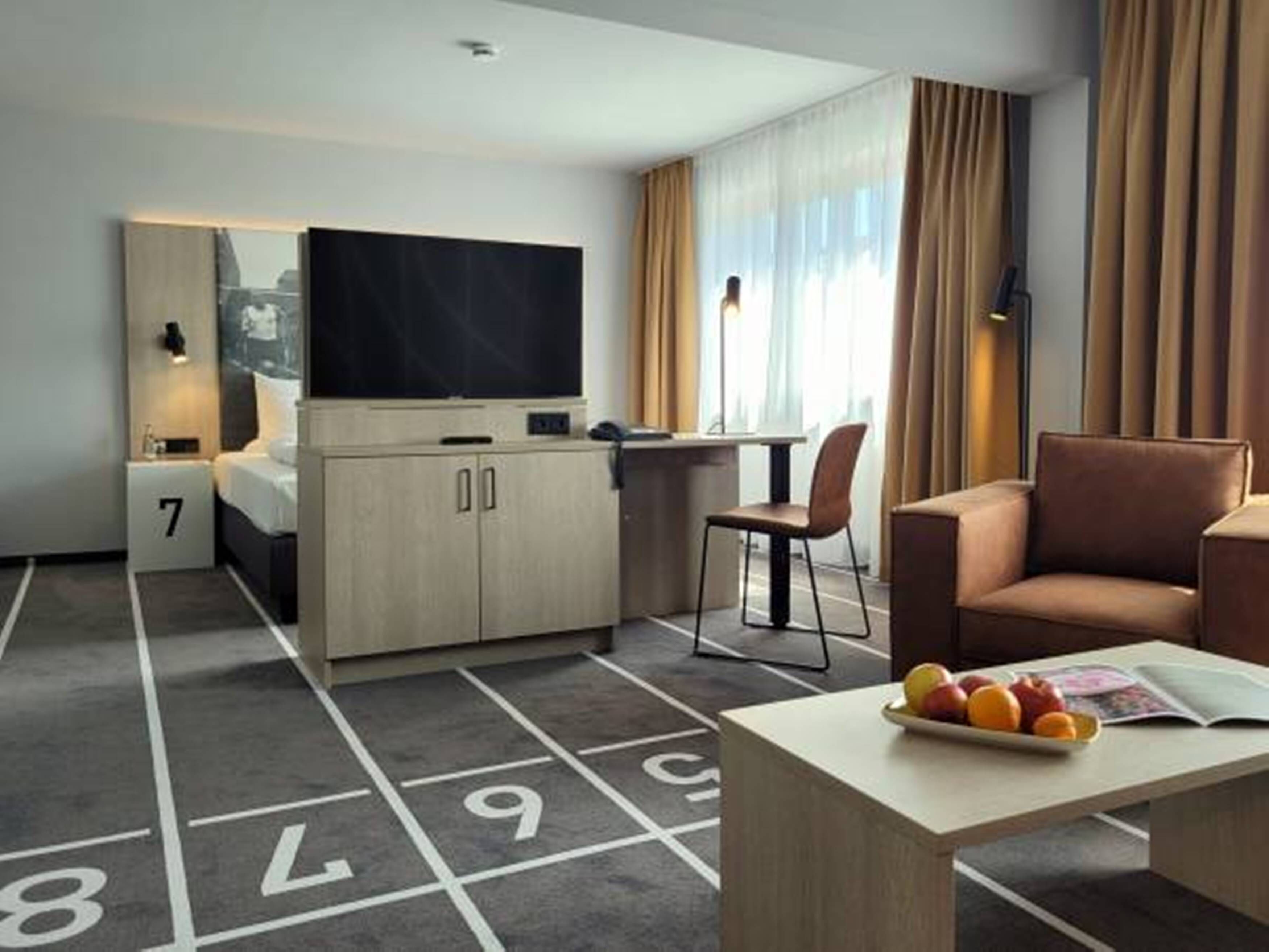 Una habitación de hotel con una cama, un escritorio, un televisor, un área de estar y una mesa de comedor con fruta.