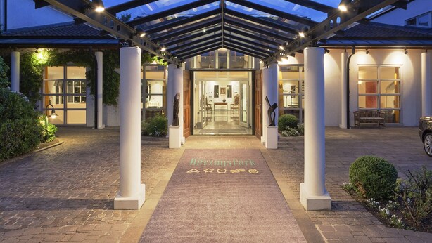 Entrada de hotel con galería techada, columnas y puerta de vidrio que da al vestíbulo.